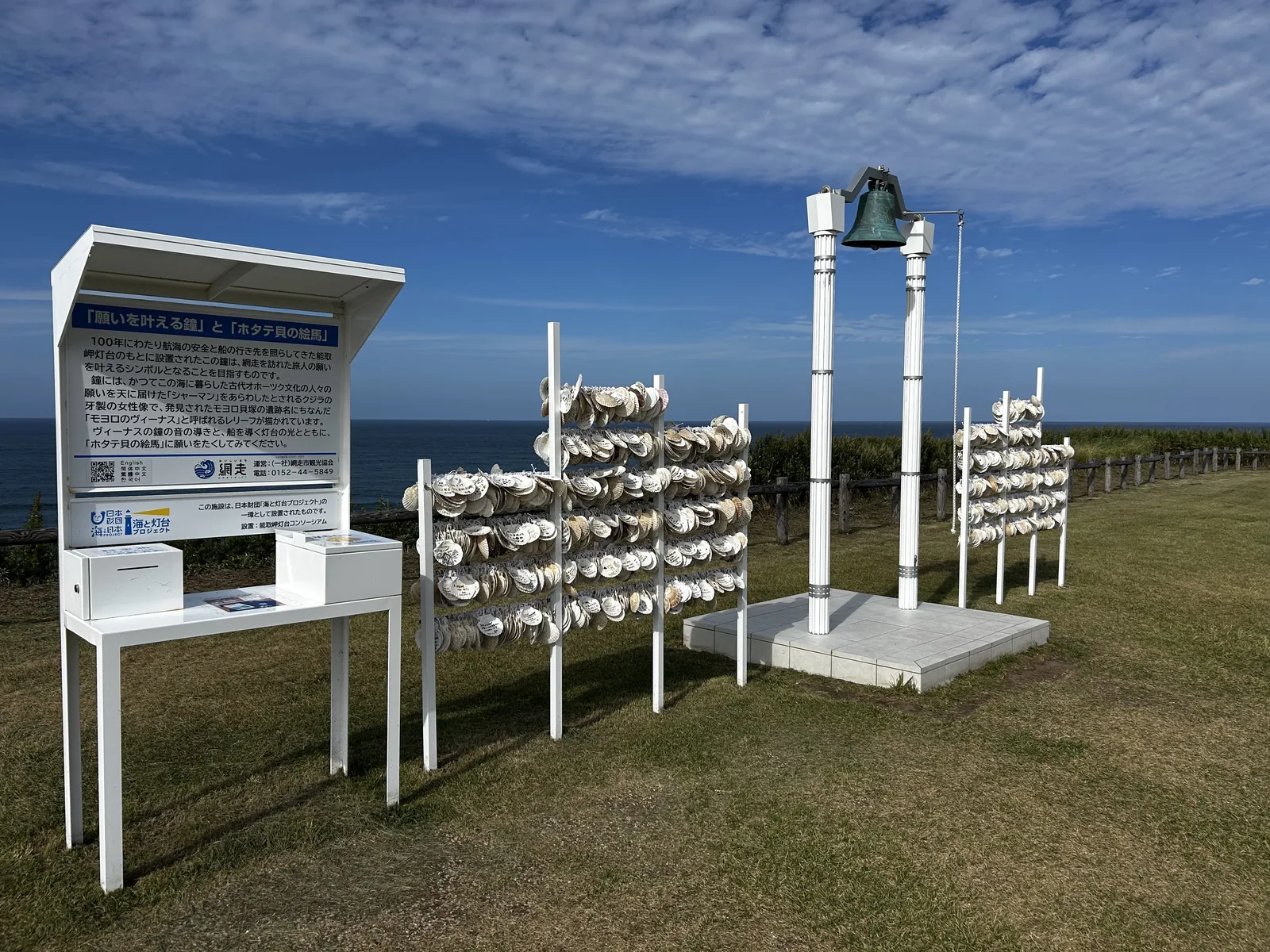 海を背景に立つ願いを叶える鐘と、白い支柱に並ぶホタテ貝の絵馬